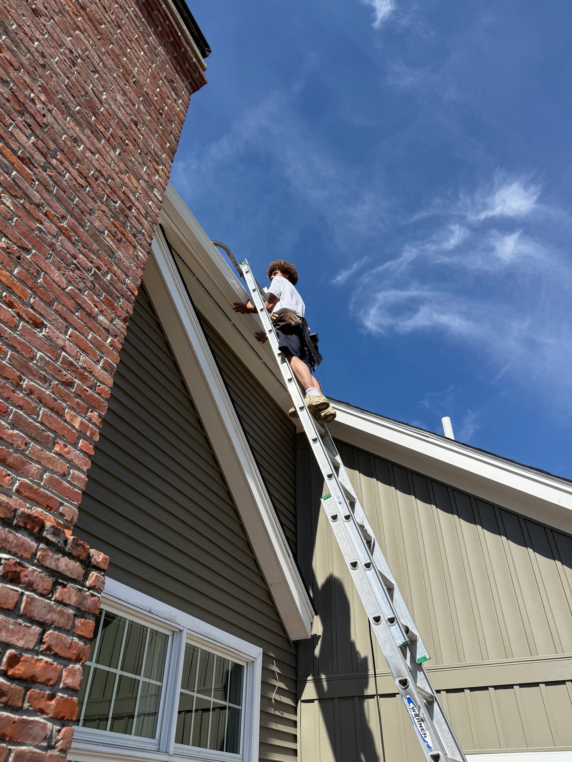 Person on a ladder cleaning a roof gutter beside a brick house