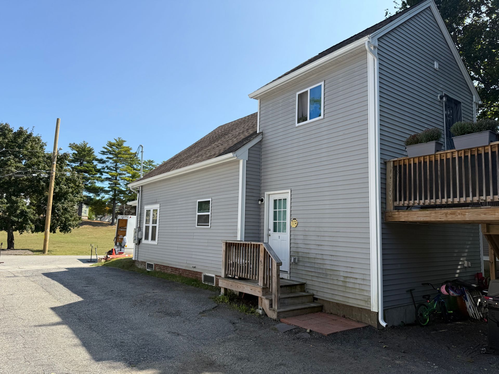 Side view of a gray house with a small porch, gravel driveway, and deck under a clear blue sky