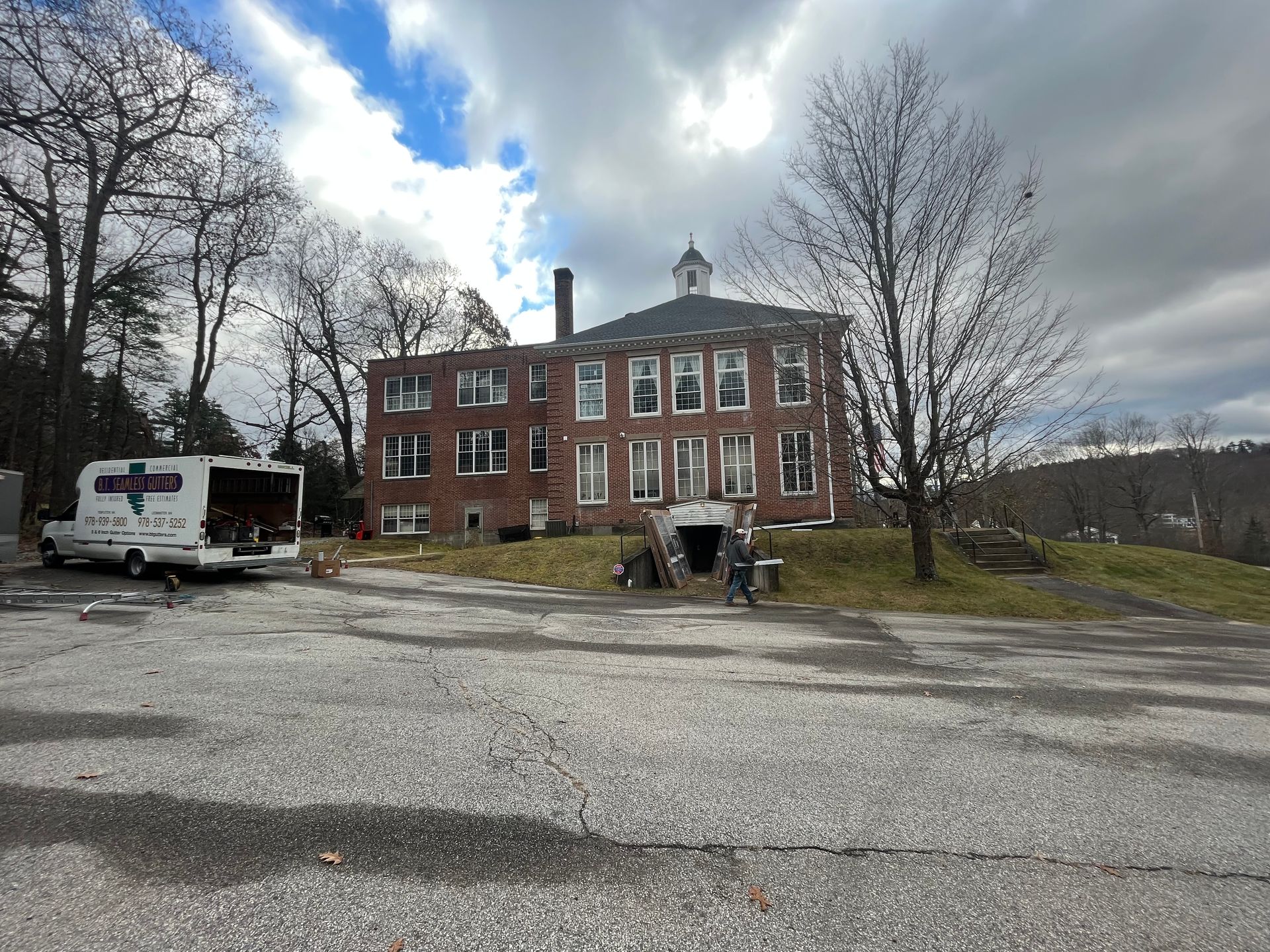 Brick building with white-trimmed windows, trees, and parked vehicles under a cloudy sky