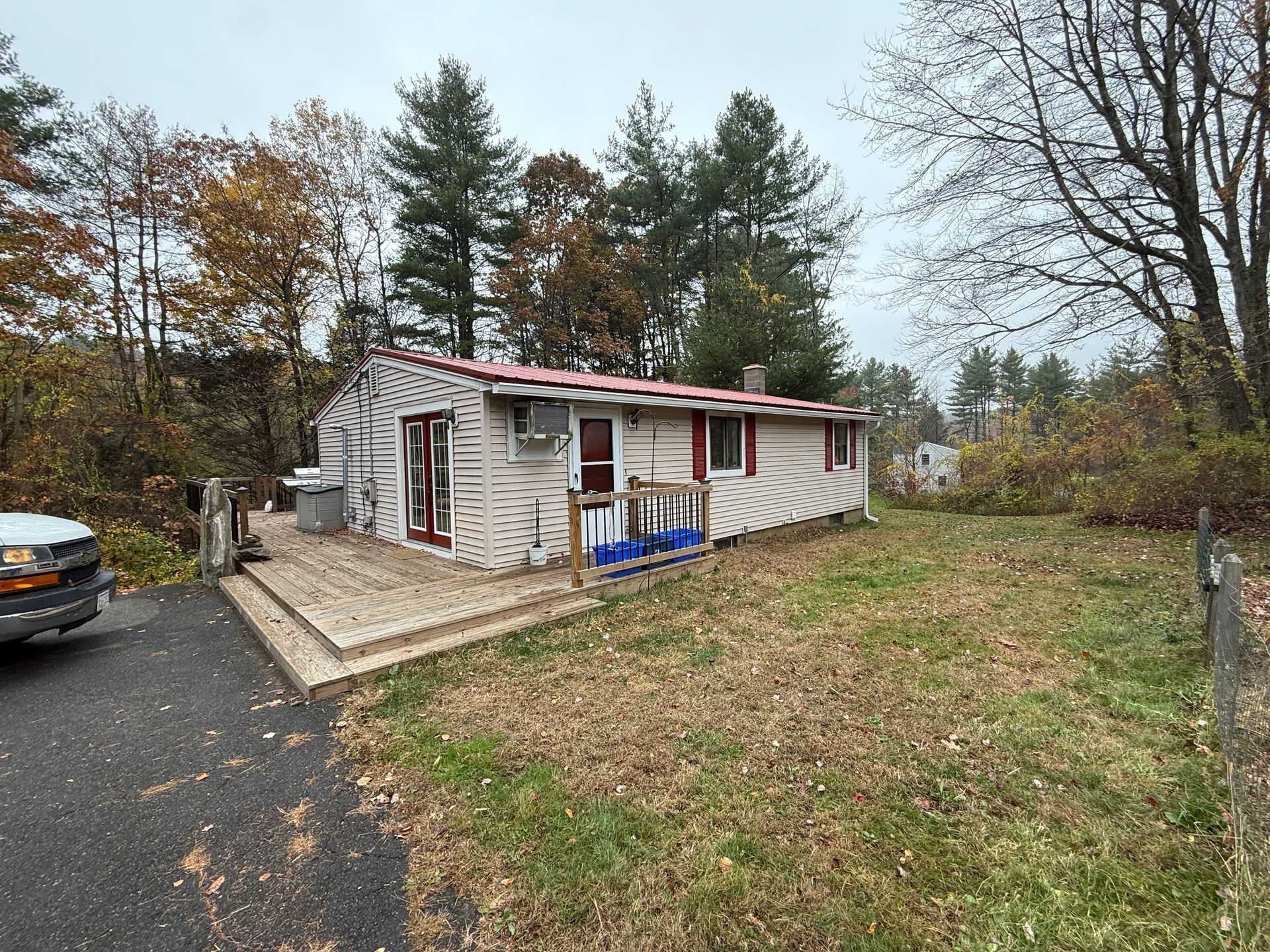 Small weathered house with a red roof beside a driveway and grassy yard in a wooded area
