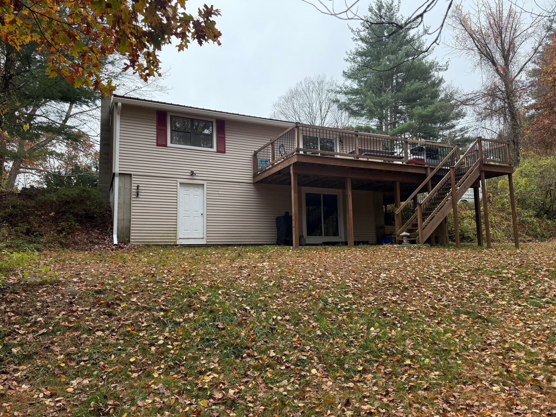 Small house with a raised wooden deck and stairs, surrounded by trees and fallen autumn leaves.