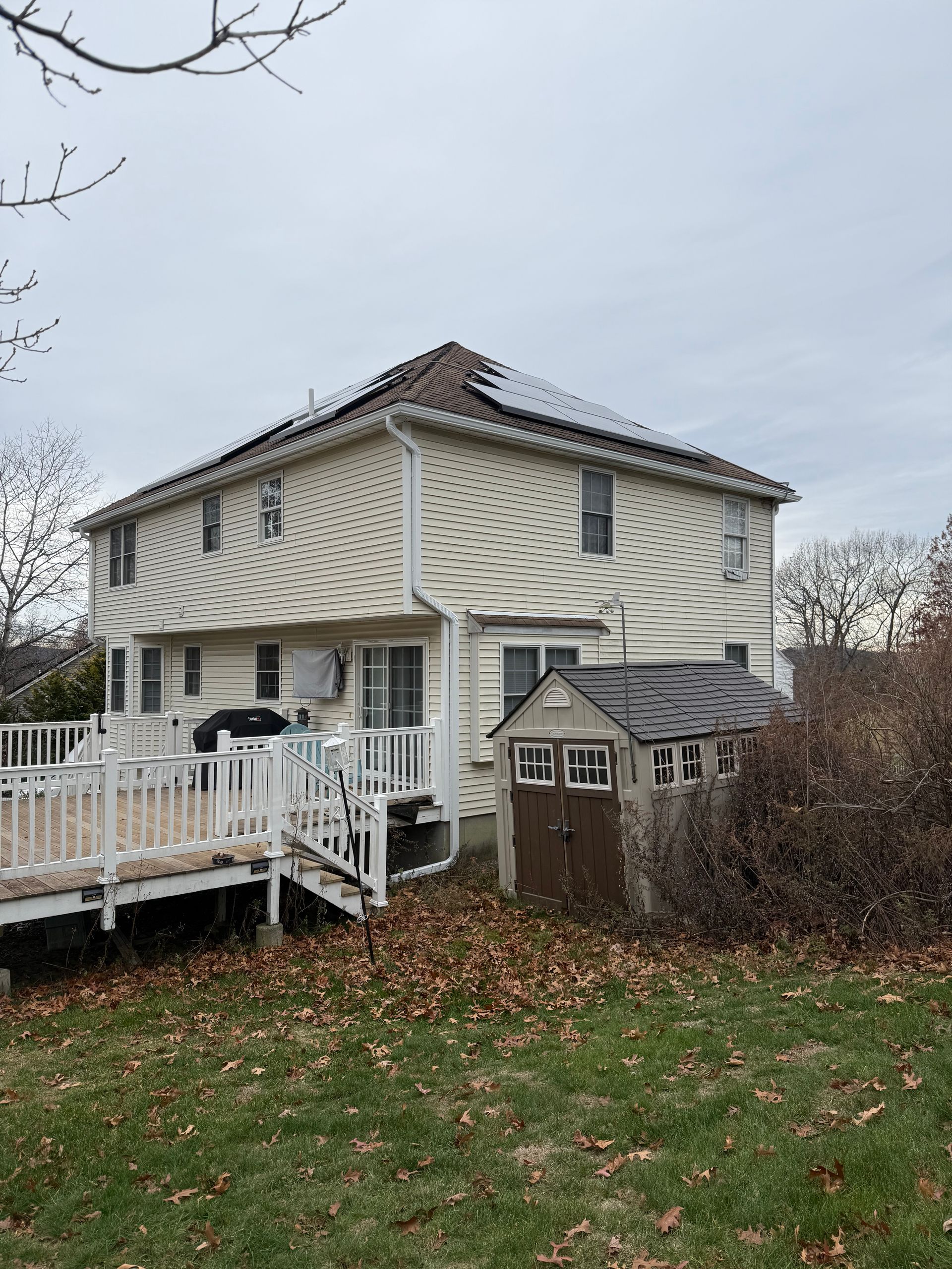 Two-story beige house with white deck and small brown shed, surrounded by fallen leaves under an overcast sky