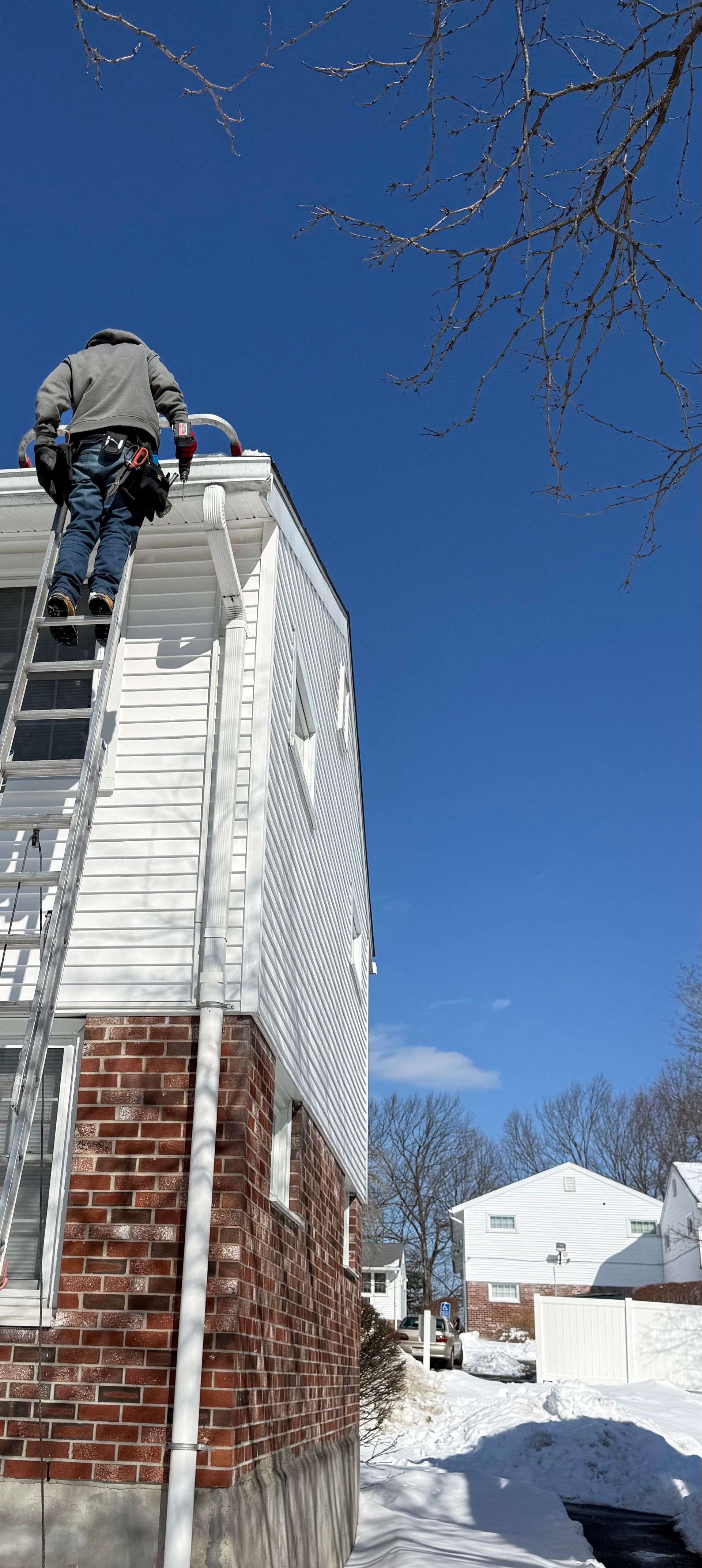 Person on roof cleaning a snow-covered white house with a red brick chimney in winter.