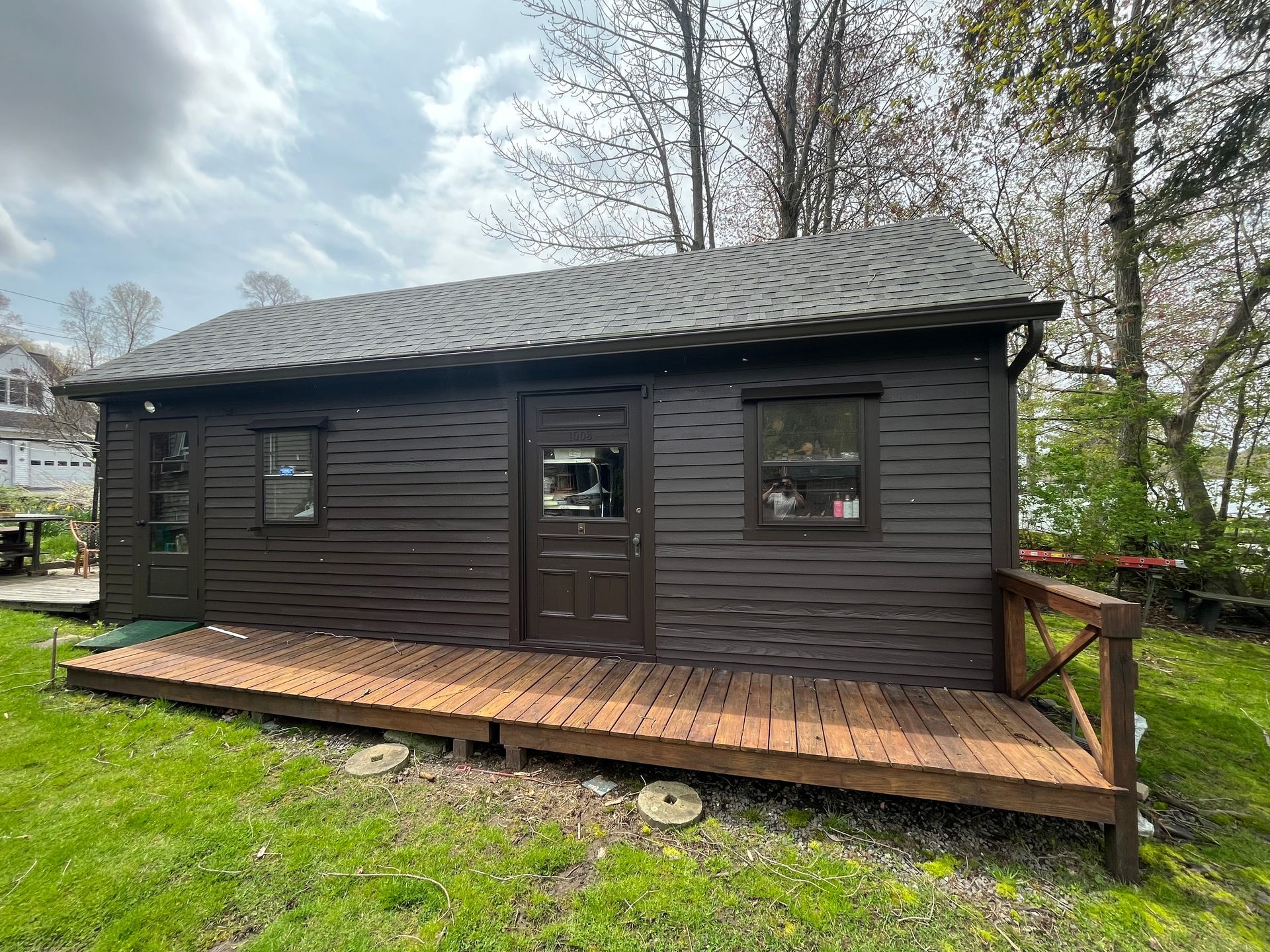 Small dark wooden cabin with a porch and metal roof in a grassy yard.