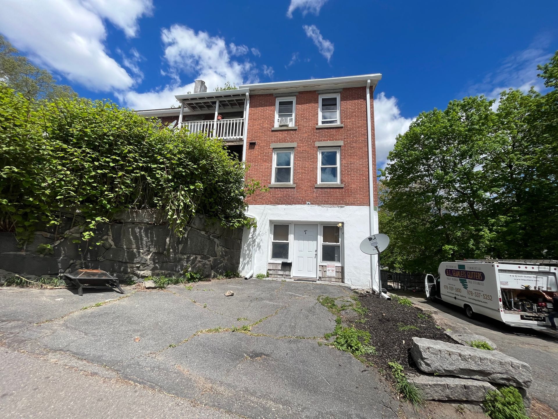 Brick building with white lower facade, gravel lot, parked vehicles, and trees under a blue sky