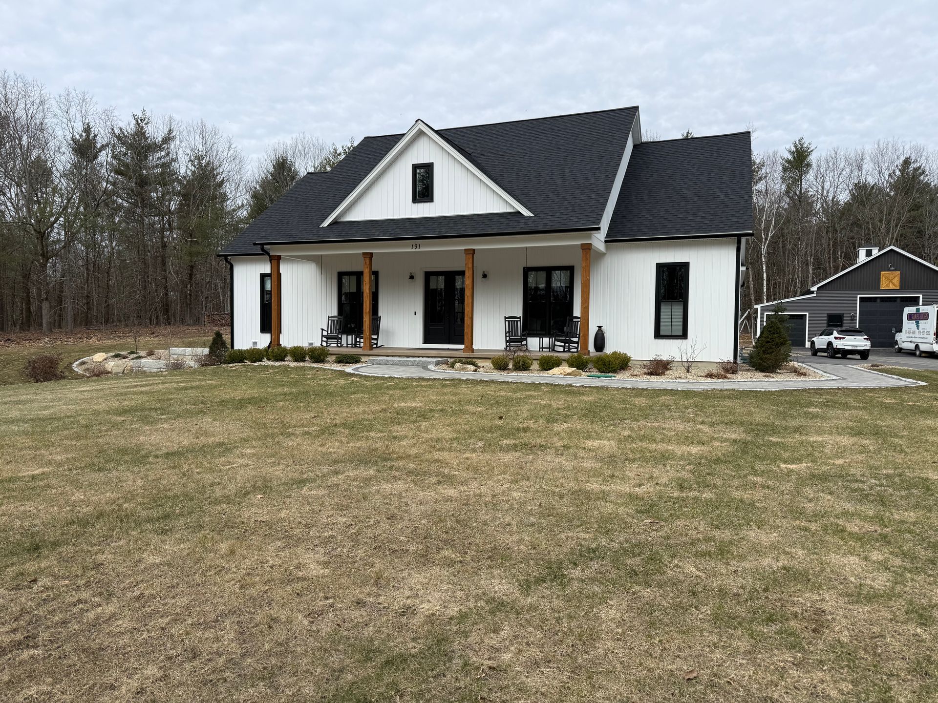 White house with black roof and porch in a grassy yard, with trees and a small outbuilding in the background