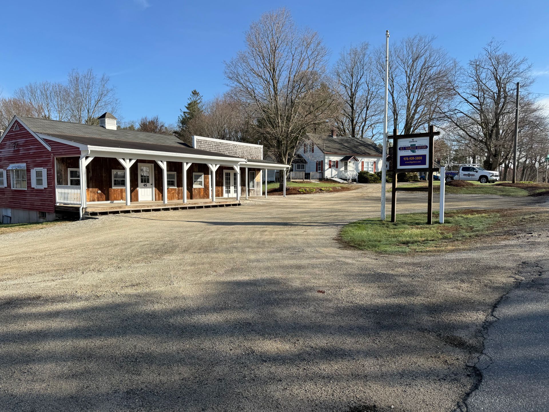 Small red-and-white building with porch beside a gravel lot and real estate sign under leafless trees