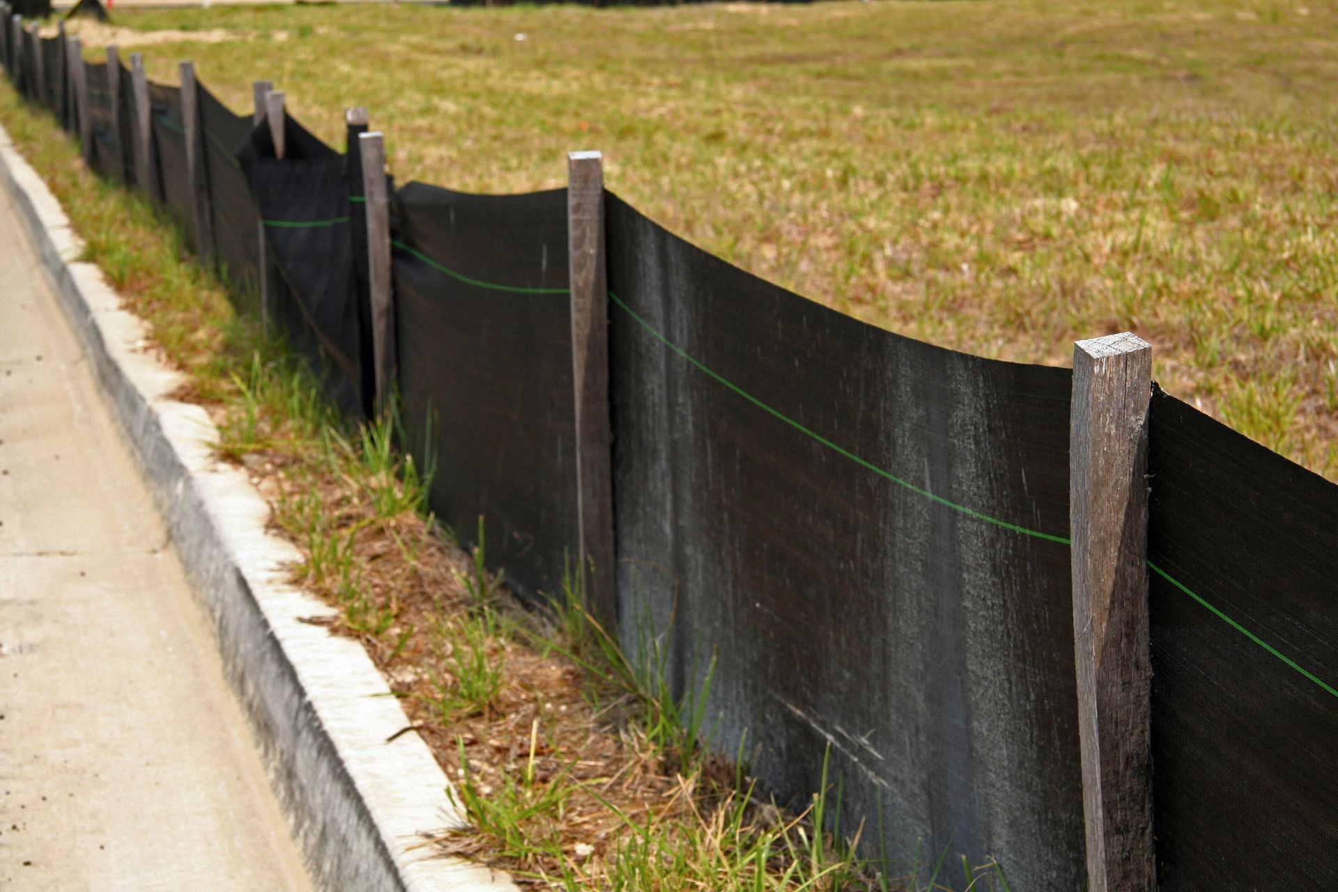 A black silt fence supported by wooden stakes runs alongside a concrete curb next to a grassy field.