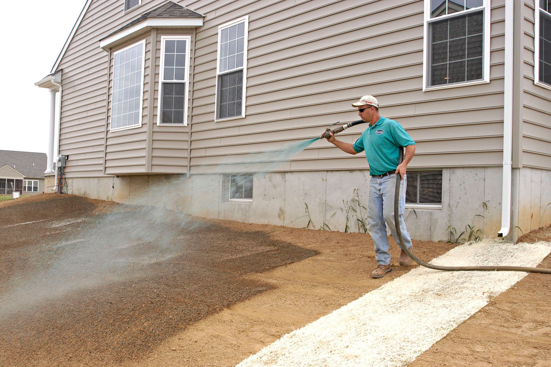 A worker in a green shirt sprays mulch onto the ground next to the concrete foundation of a beige siding house.