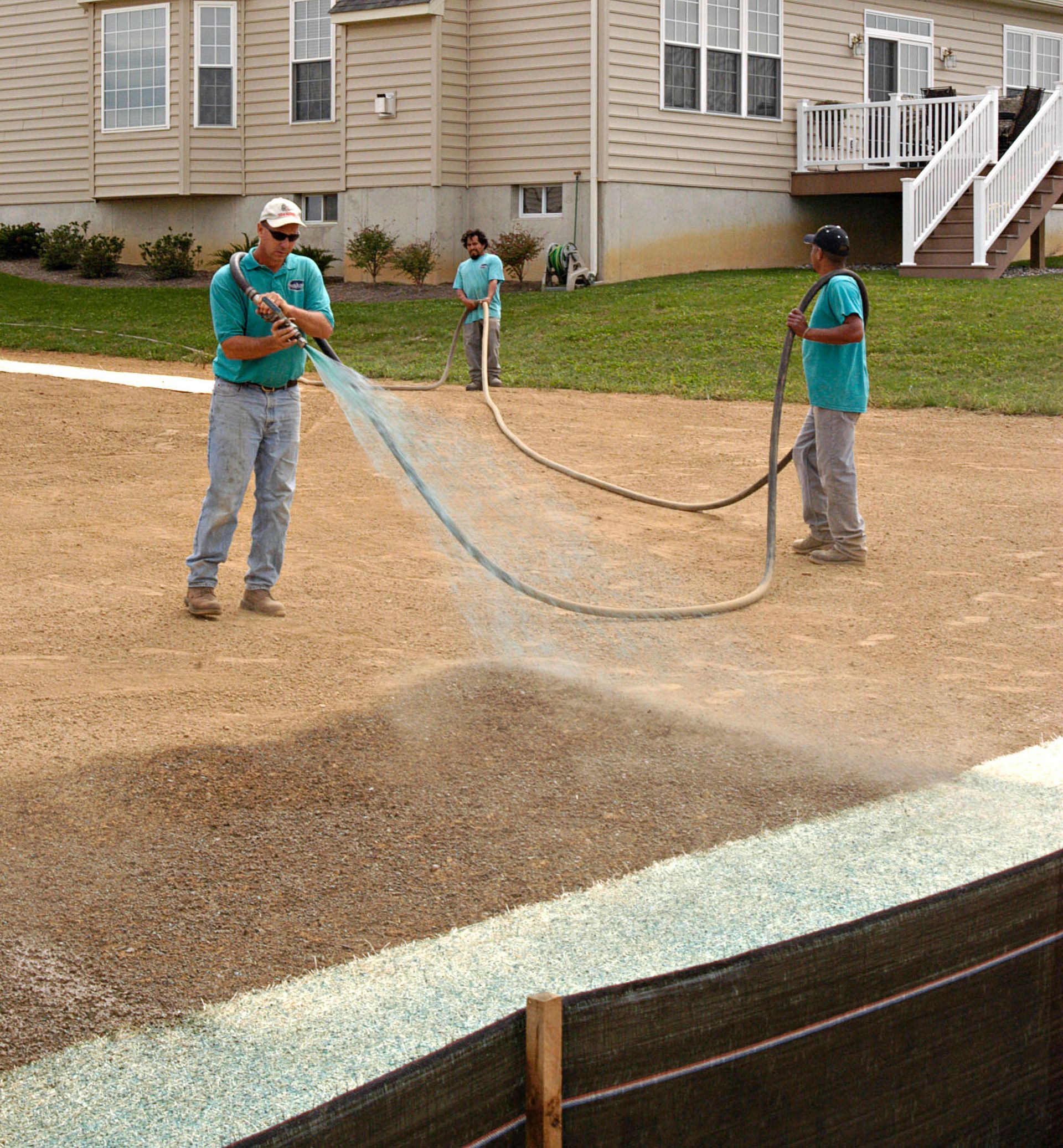 Three workers in turquoise shirts use a hose to spray a mixture onto a bare lawn in front of a residential house.