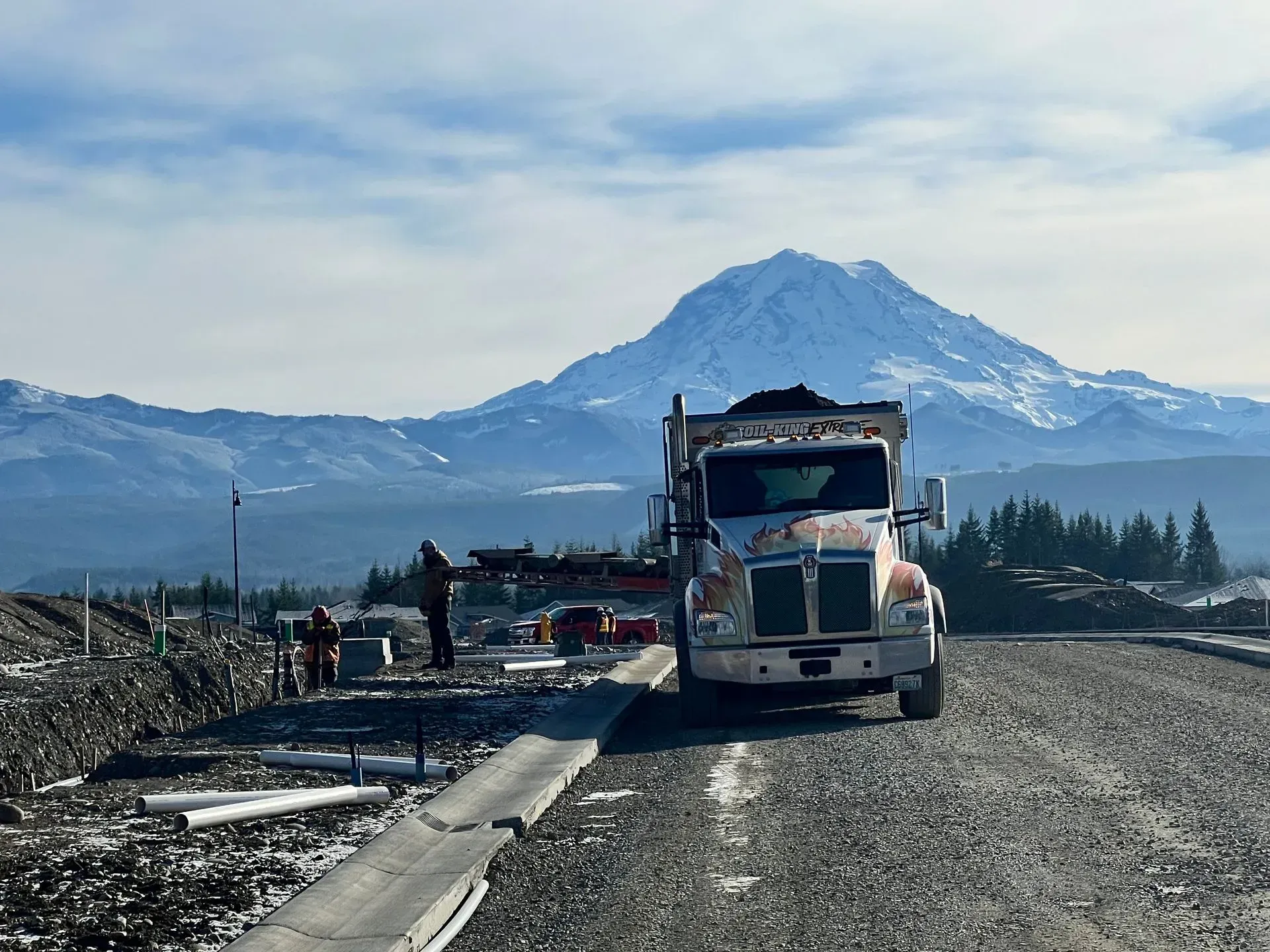 A construction dump truck sits on a gravel road with a large, snow-capped mountain in the background.