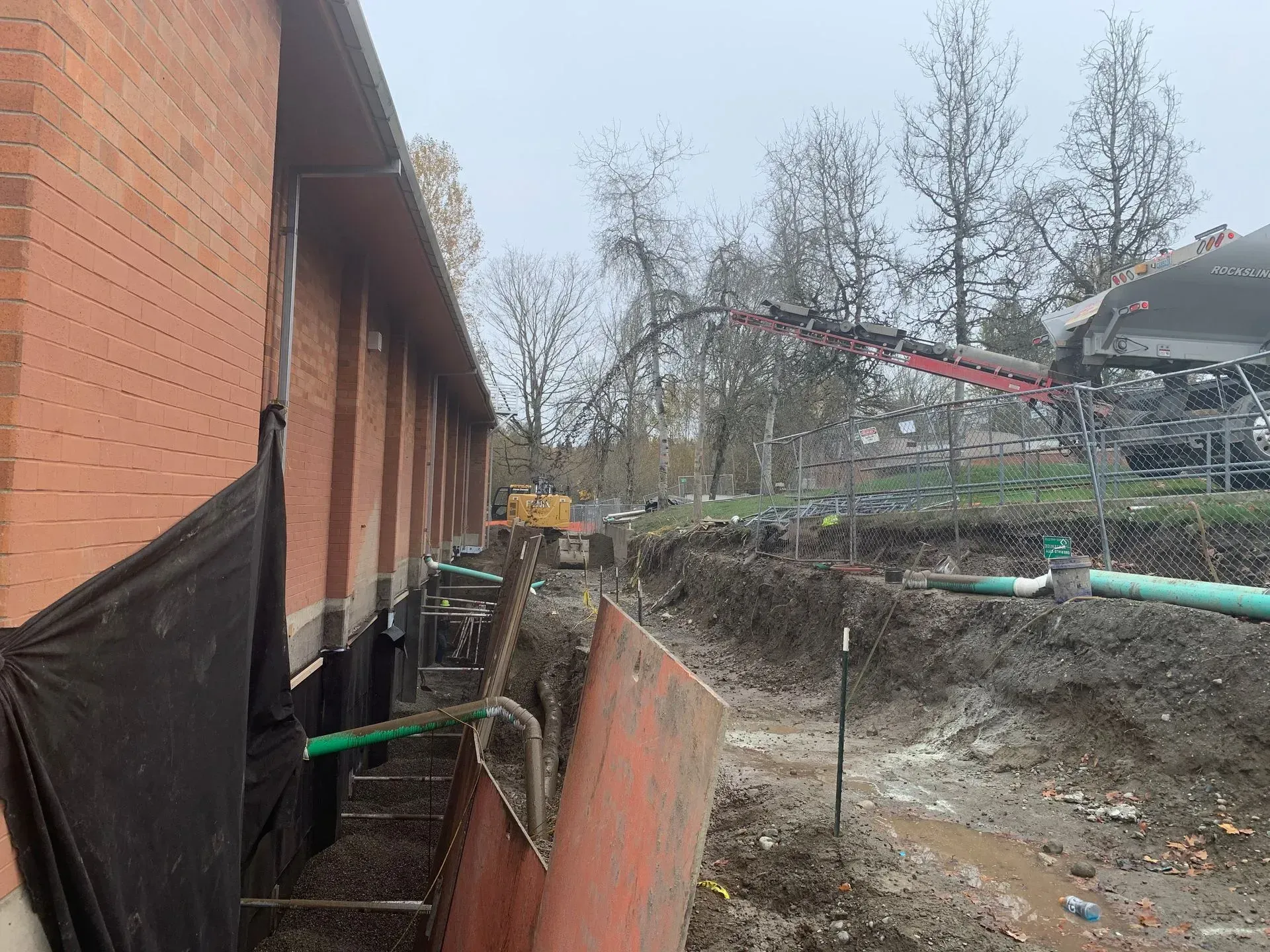 Construction site showing a brick building foundation, exposed soil, and machinery for earthwork and pipe installation.
