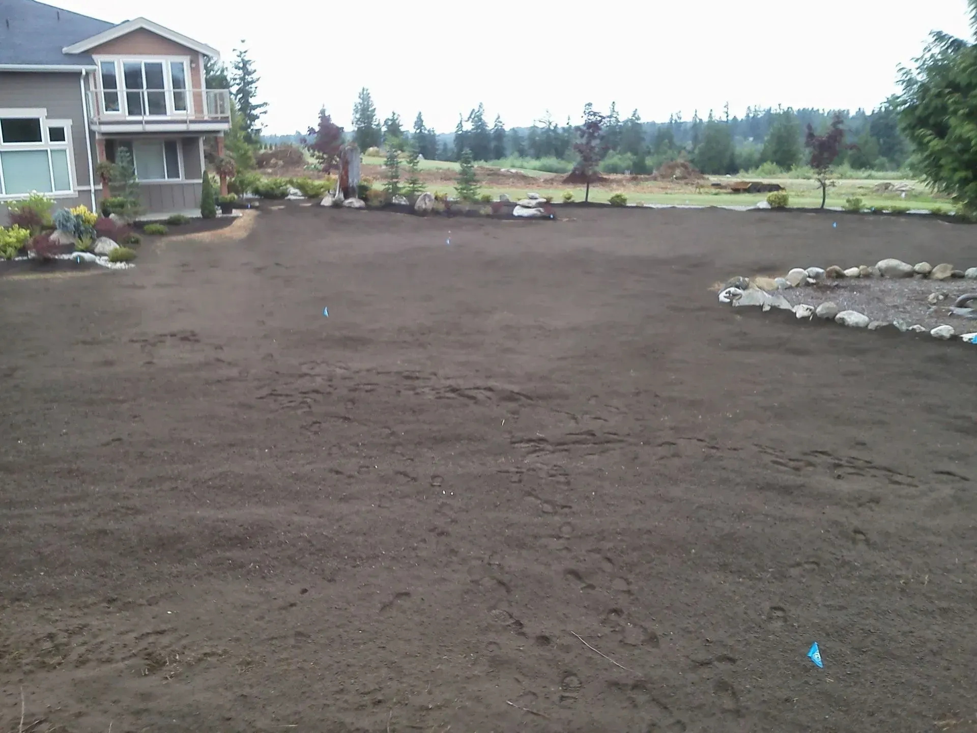 A suburban house with a large, freshly graded, bare dirt yard and a stone garden border on a cloudy day.