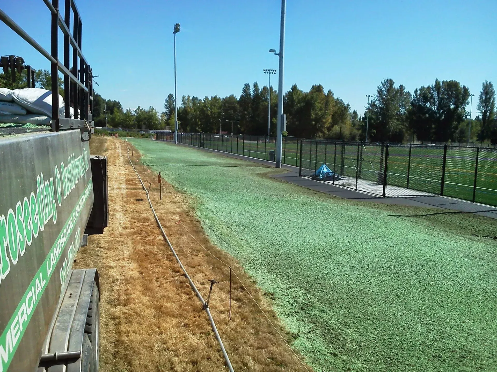 A strip of bare dirt beside a lush green turf field, separated by a fence with stadium lights under a clear blue sky.