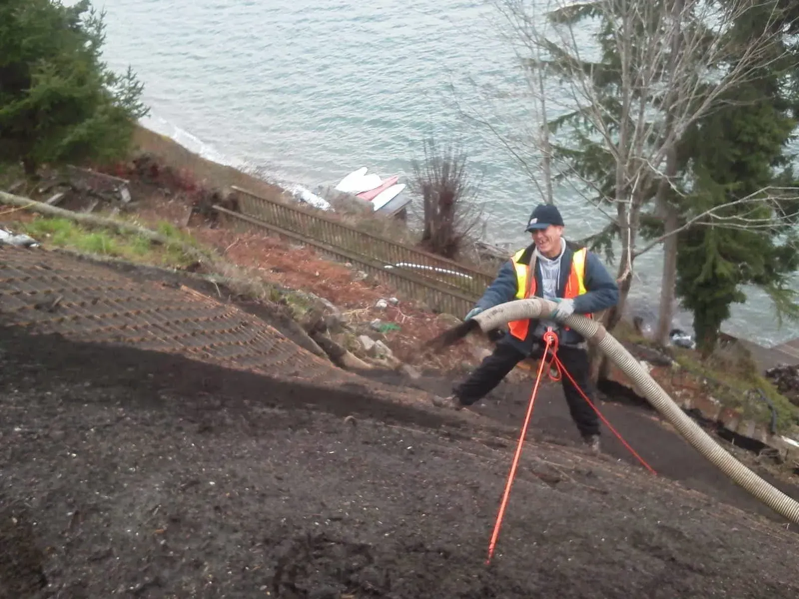 A person in a high-visibility vest stands on a steep, dirt embankment, directing a hose to spread material onto the slope.