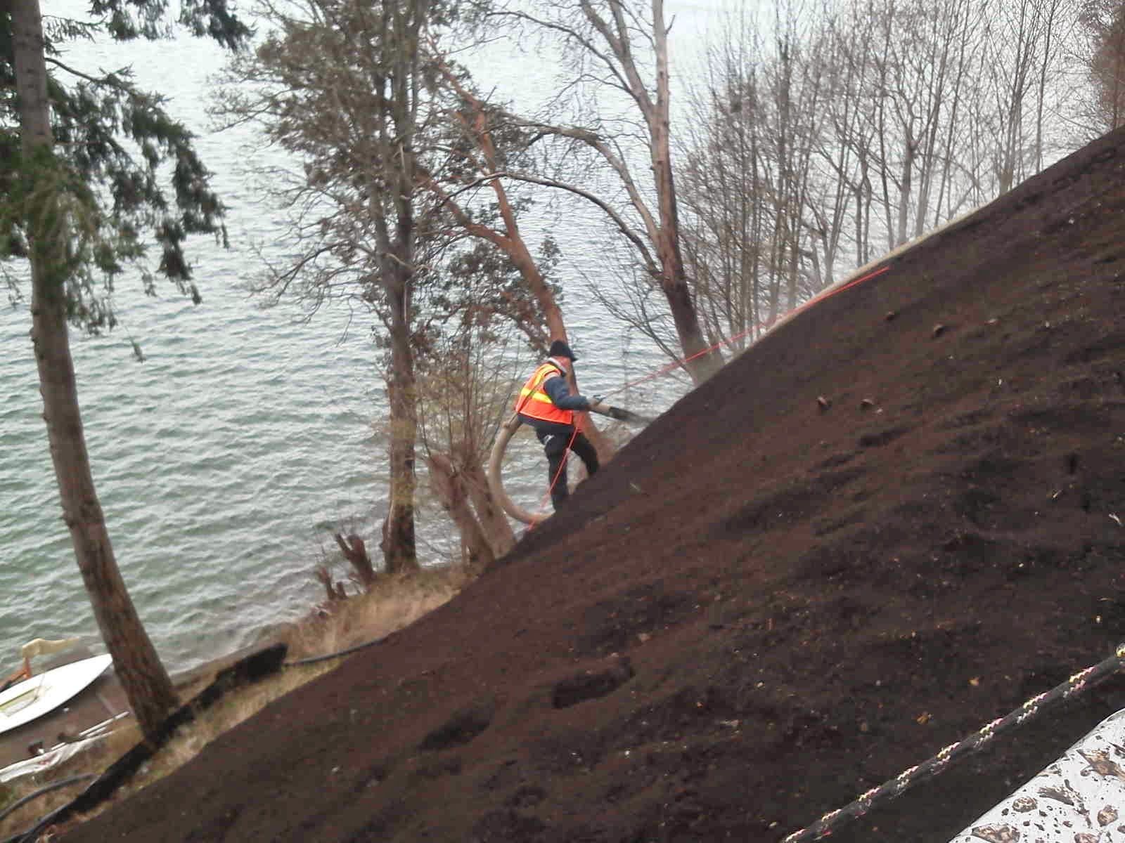A worker in a high-visibility vest sprays dark material onto a steep, bare embankment next to a body of water.