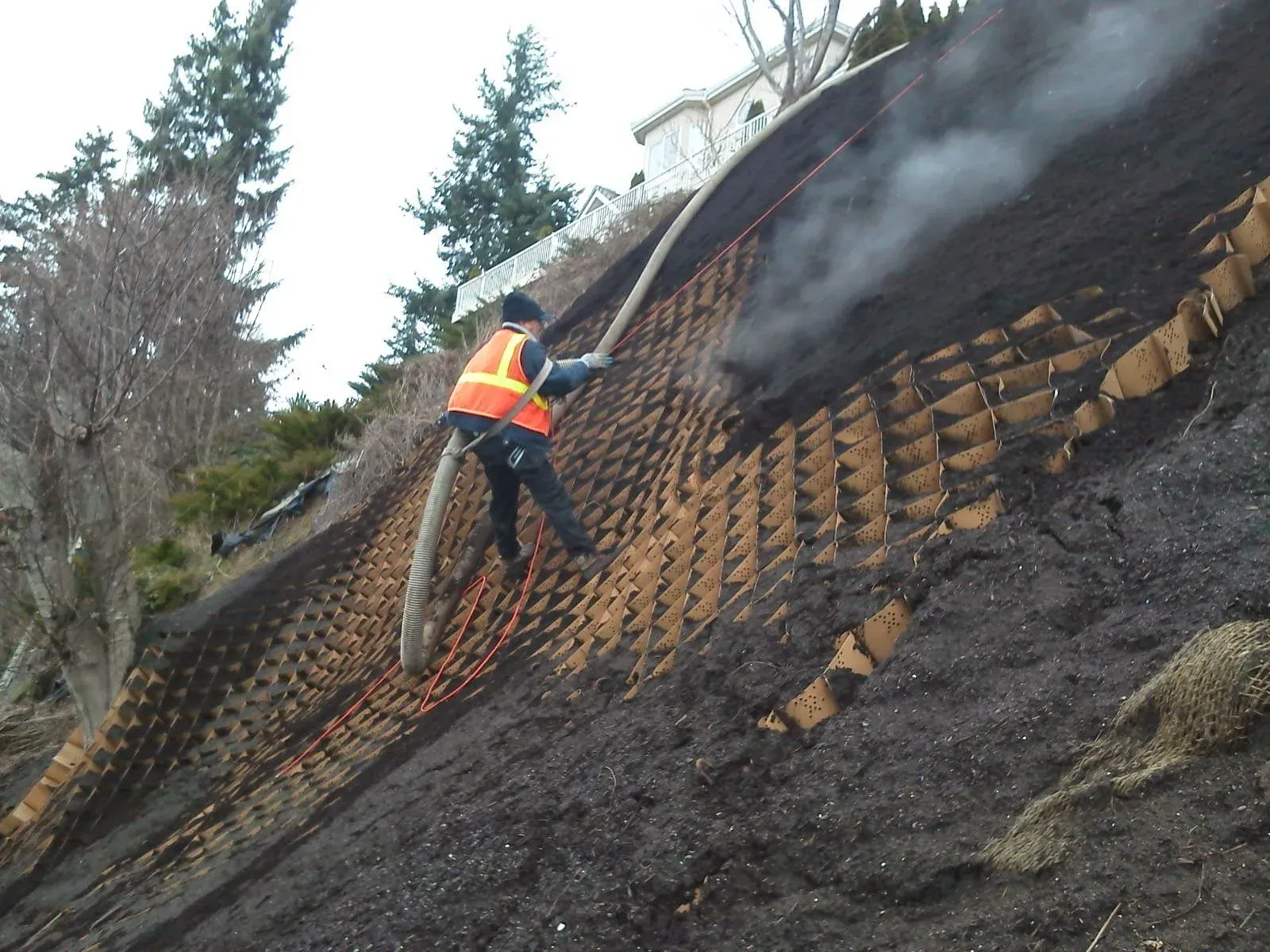 A worker in an orange safety vest sprays mulch onto a sloped embankment reinforced with a brown, honeycomb grid structure.