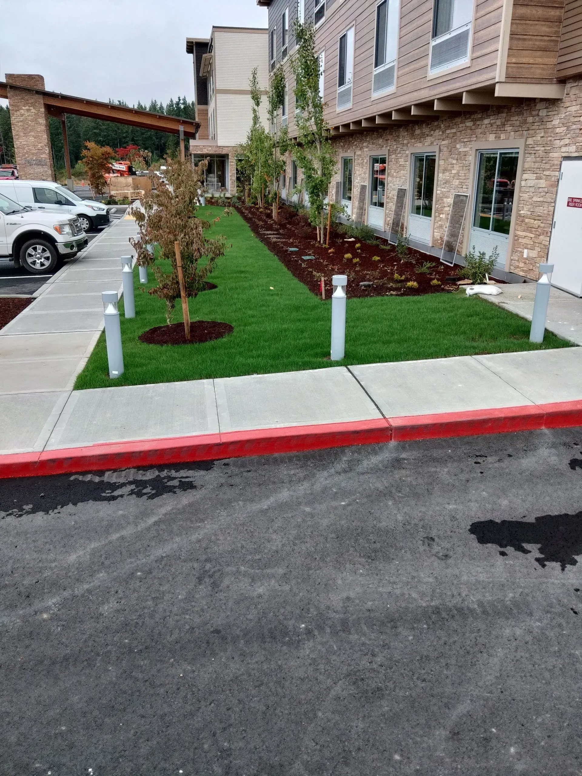 A view of a hotel building exterior with a green lawn, young trees, and a red curb alongside an asphalt parking lot.