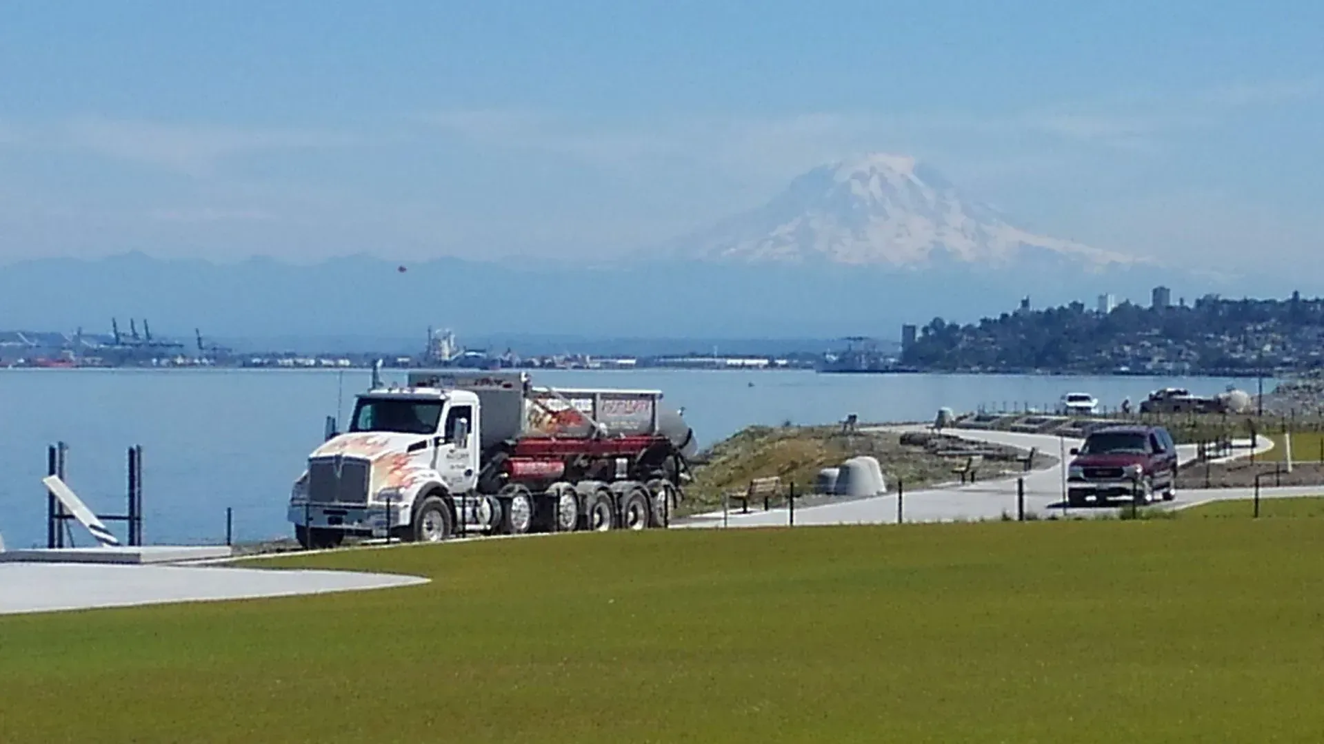 A white dump truck sits on a waterfront path with Mount Rainier in the background.