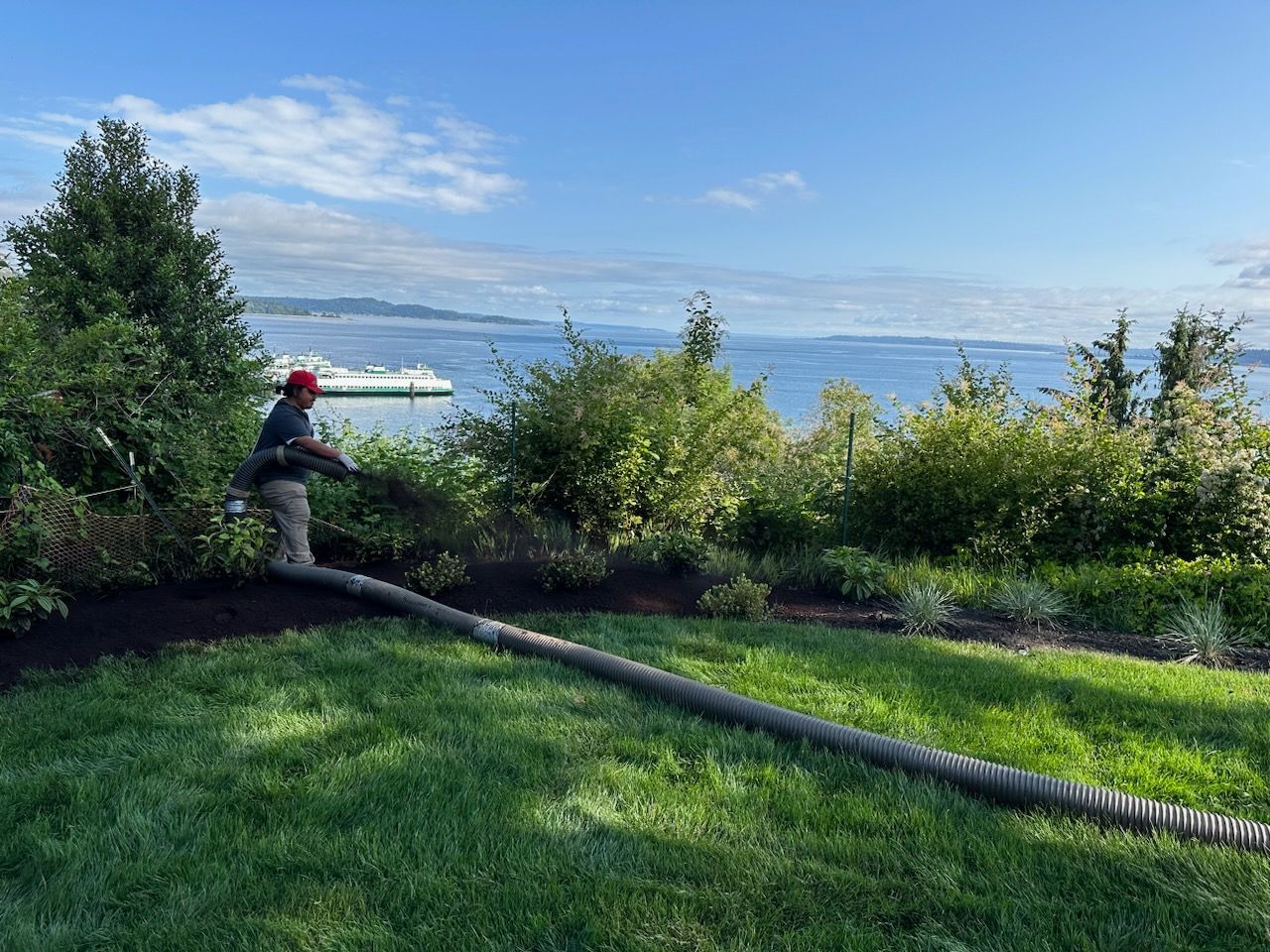 Person in a red hat standing by a hose on a grassy lawn overlooking a lake and distant mountains.