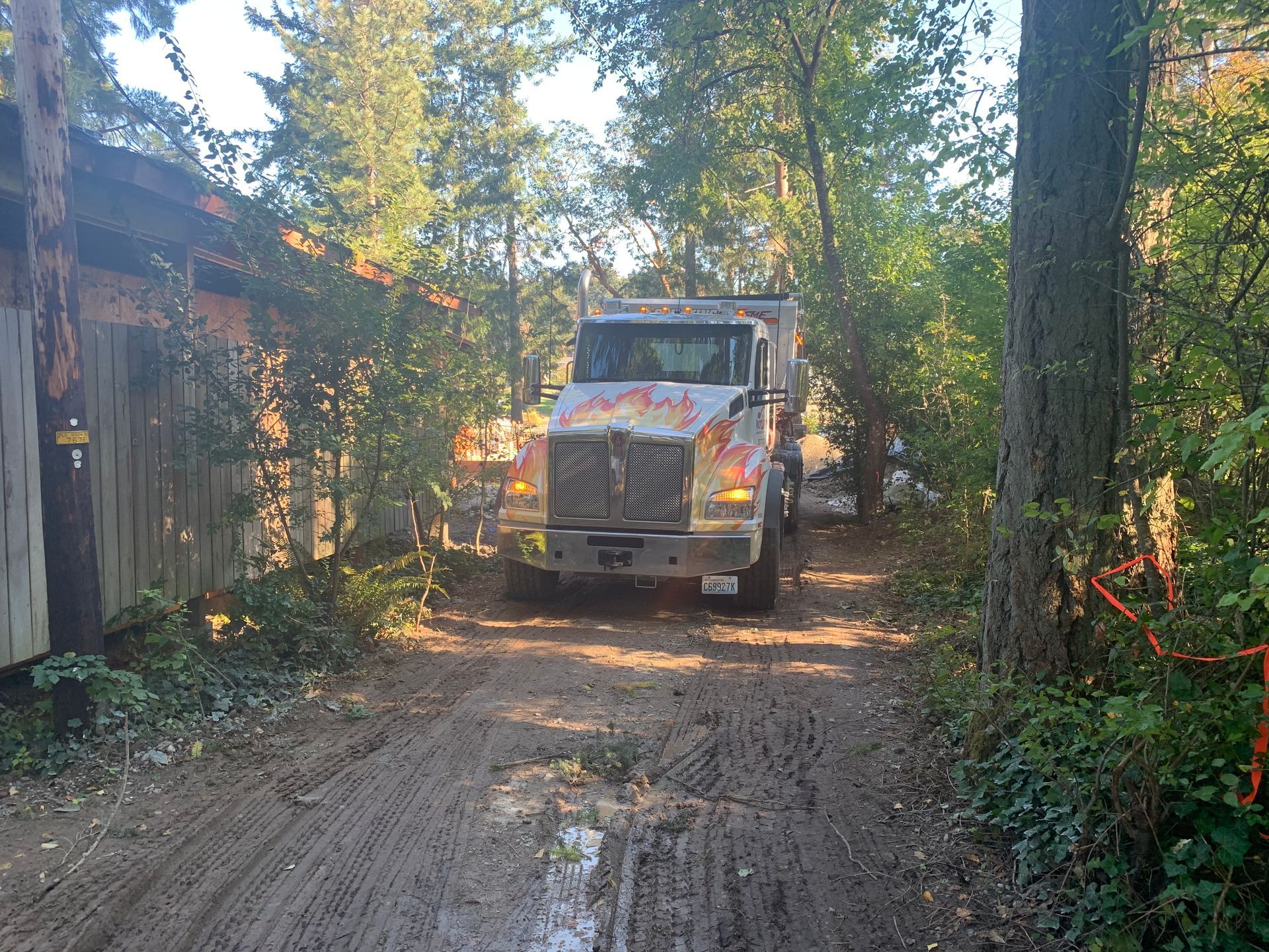 Dump truck on a muddy forest road beside a wooden building, with trees and sunlight in the background.