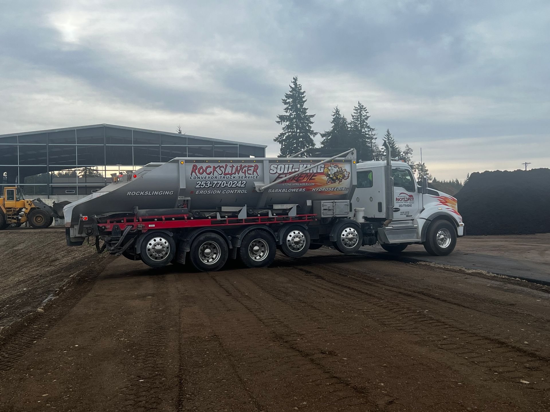 White dump truck with red trailer driving on a dirt track beside a stadium at dusk