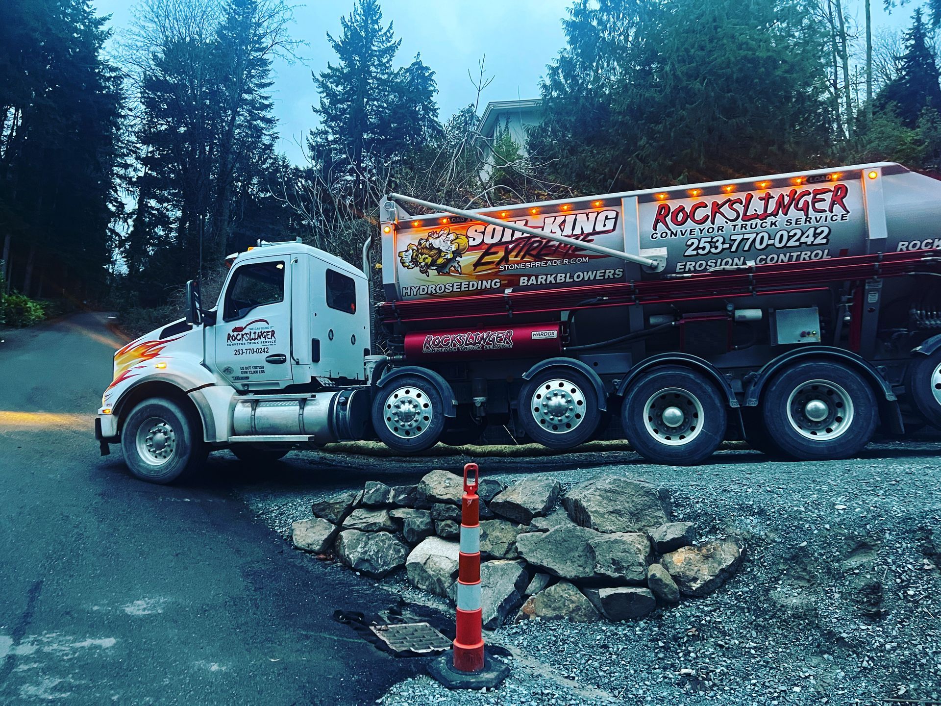 White tow truck with red trailer on a rocky roadside in a wooded area, with orange traffic cones nearby