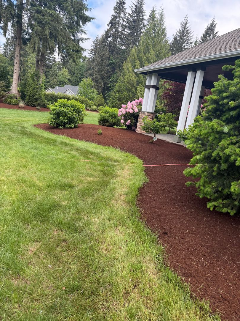 Landscaped front yard with green lawn, mulch border, shrubs, and a house porch with white columns.