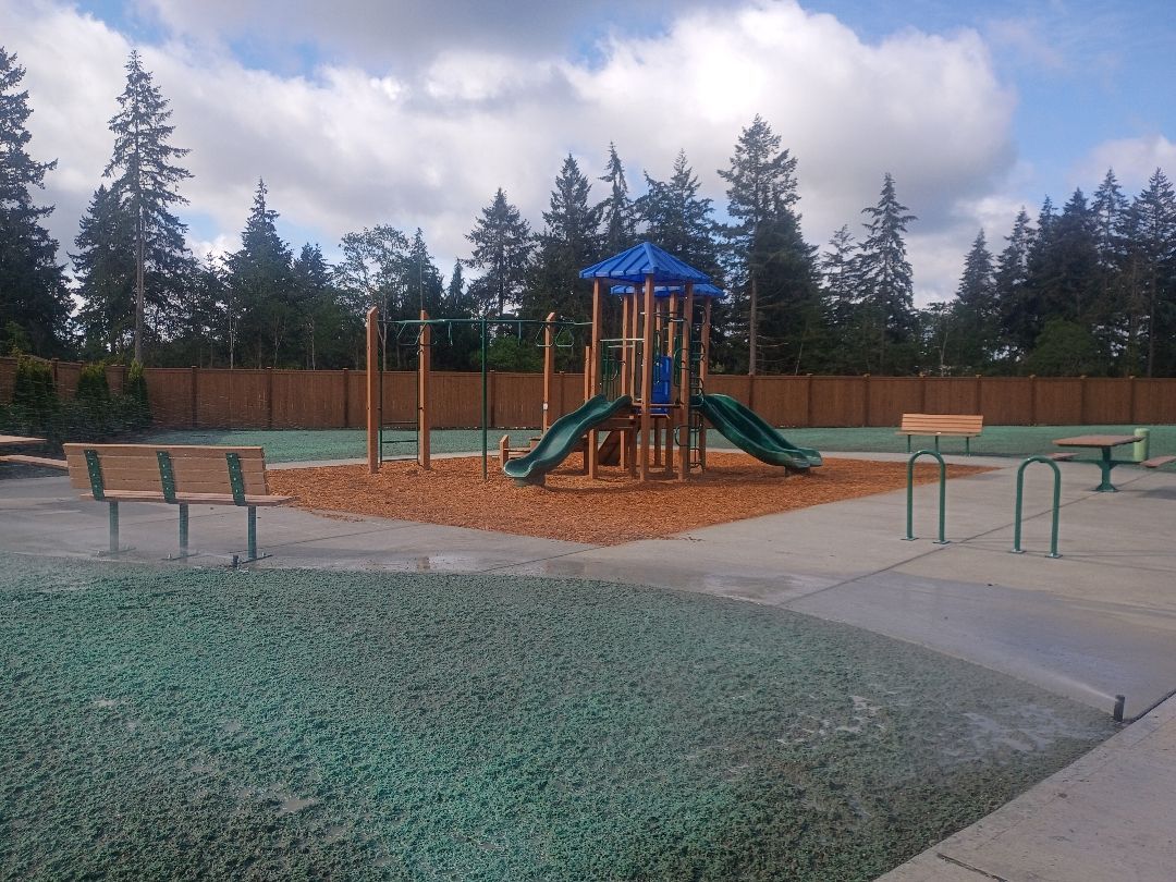 Empty playground with slides, benches, and wood chips under a cloudy sky, surrounded by trees.
