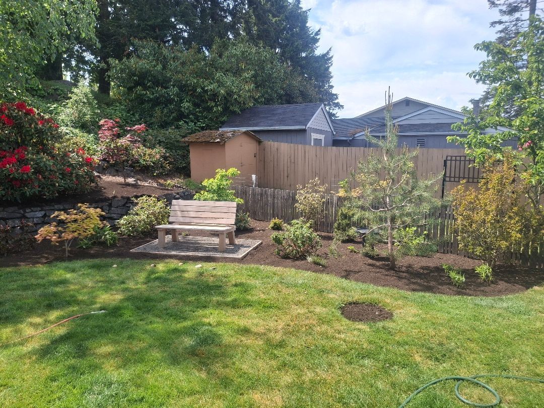 A backyard garden with lawn, flower beds, a bench, and a wooden fence under trees
