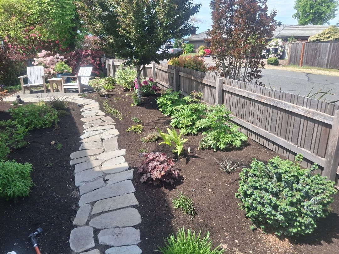 Garden path with stone pavers beside mulched flower beds, shrubs, and a wooden fence on a sunny day