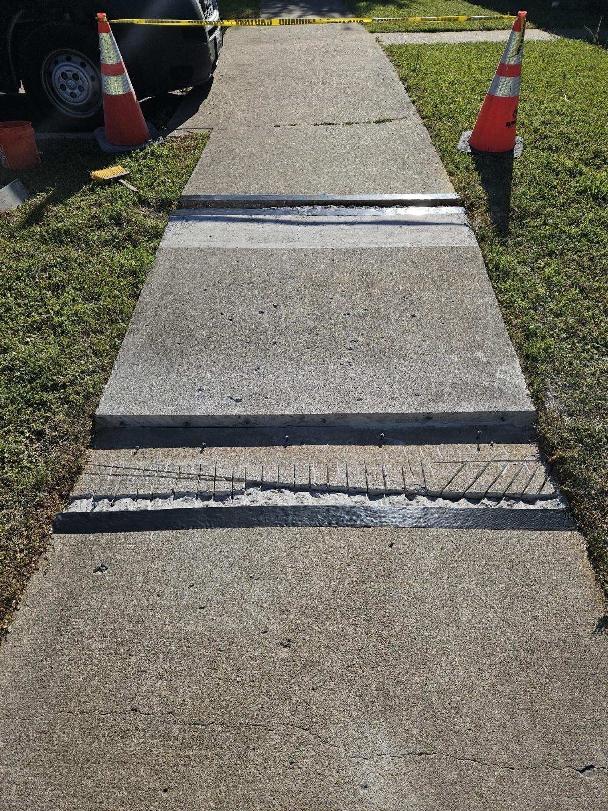 Sidewalk with uneven sections, concrete and black sealant. Orange traffic cones on each side.