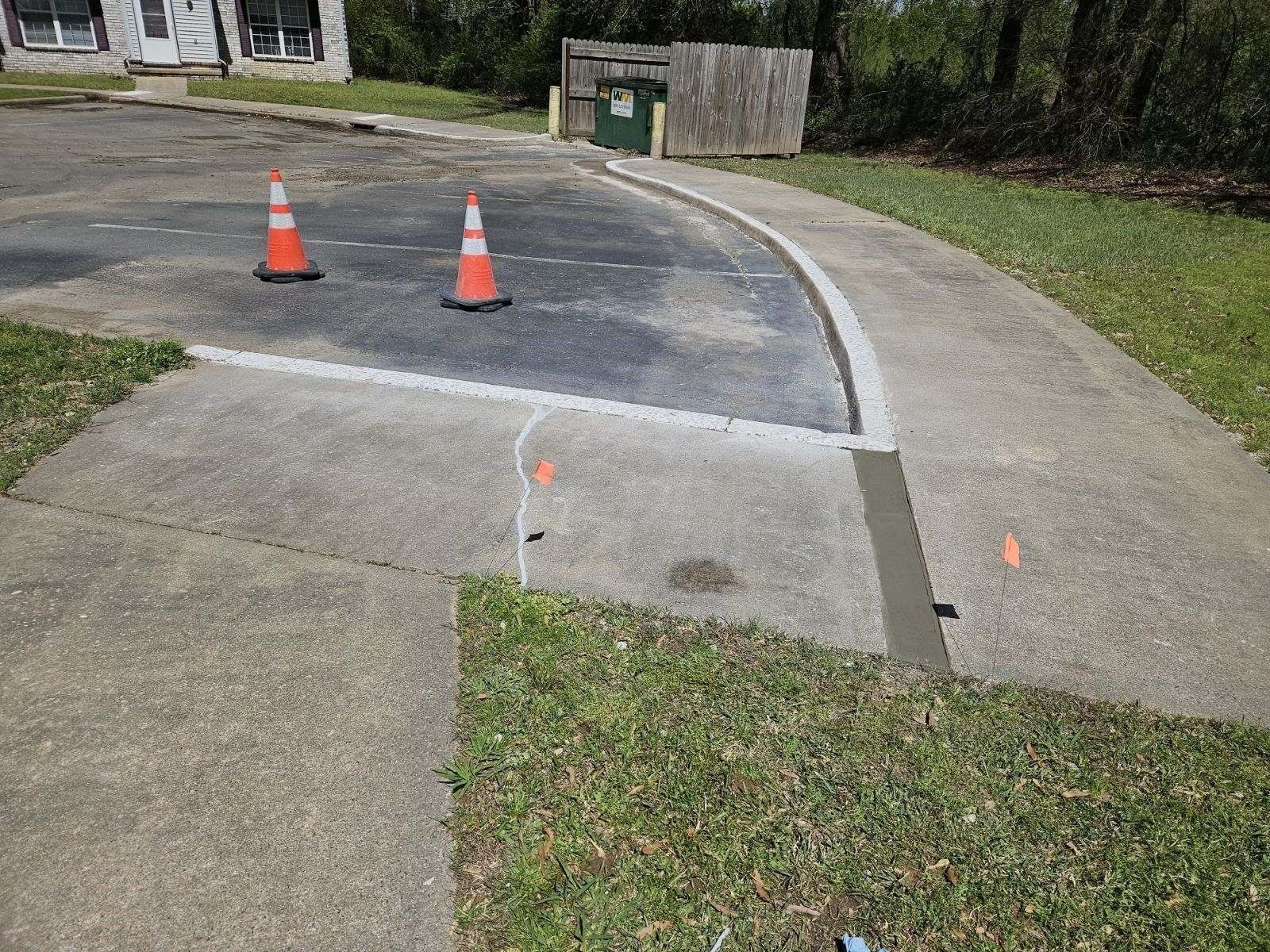 Concrete sidewalk and driveway with two orange cones, grass, and a dumpster.