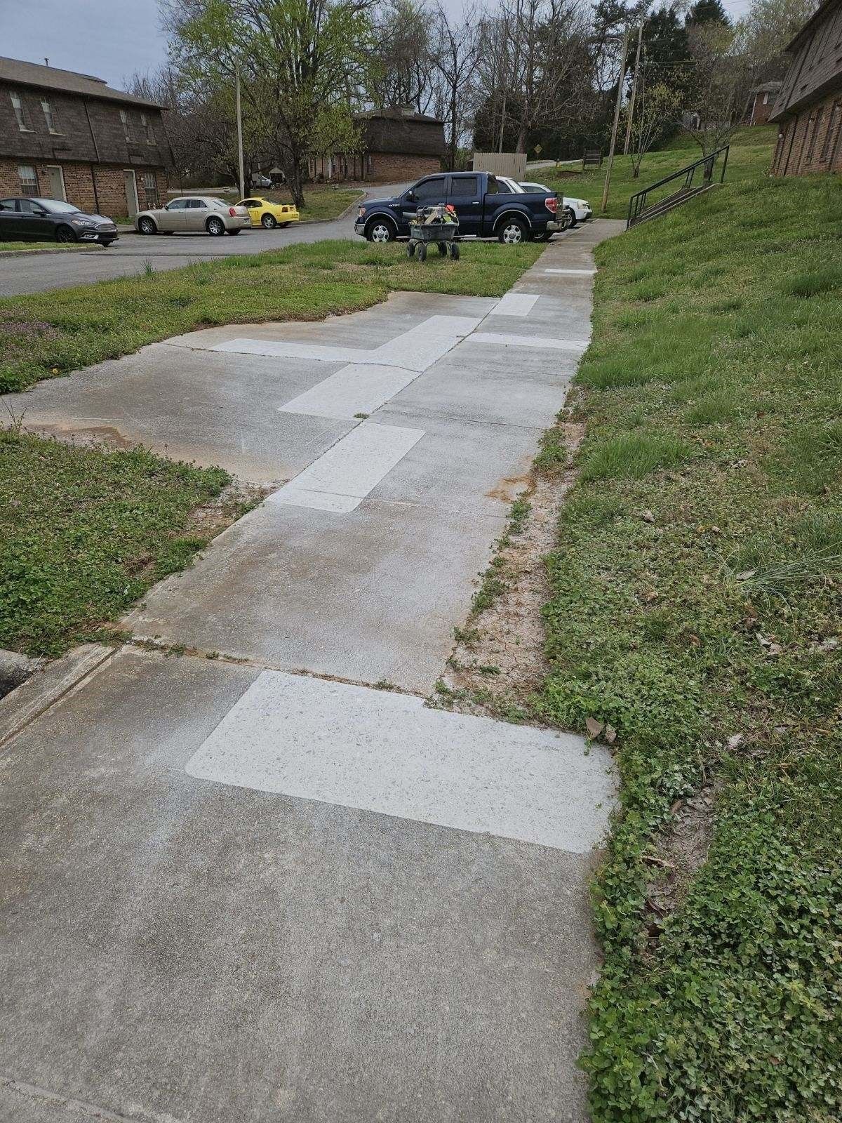 Sidewalk with concrete patches, bordered by grass, leading toward parked cars and buildings. Overcast sky.