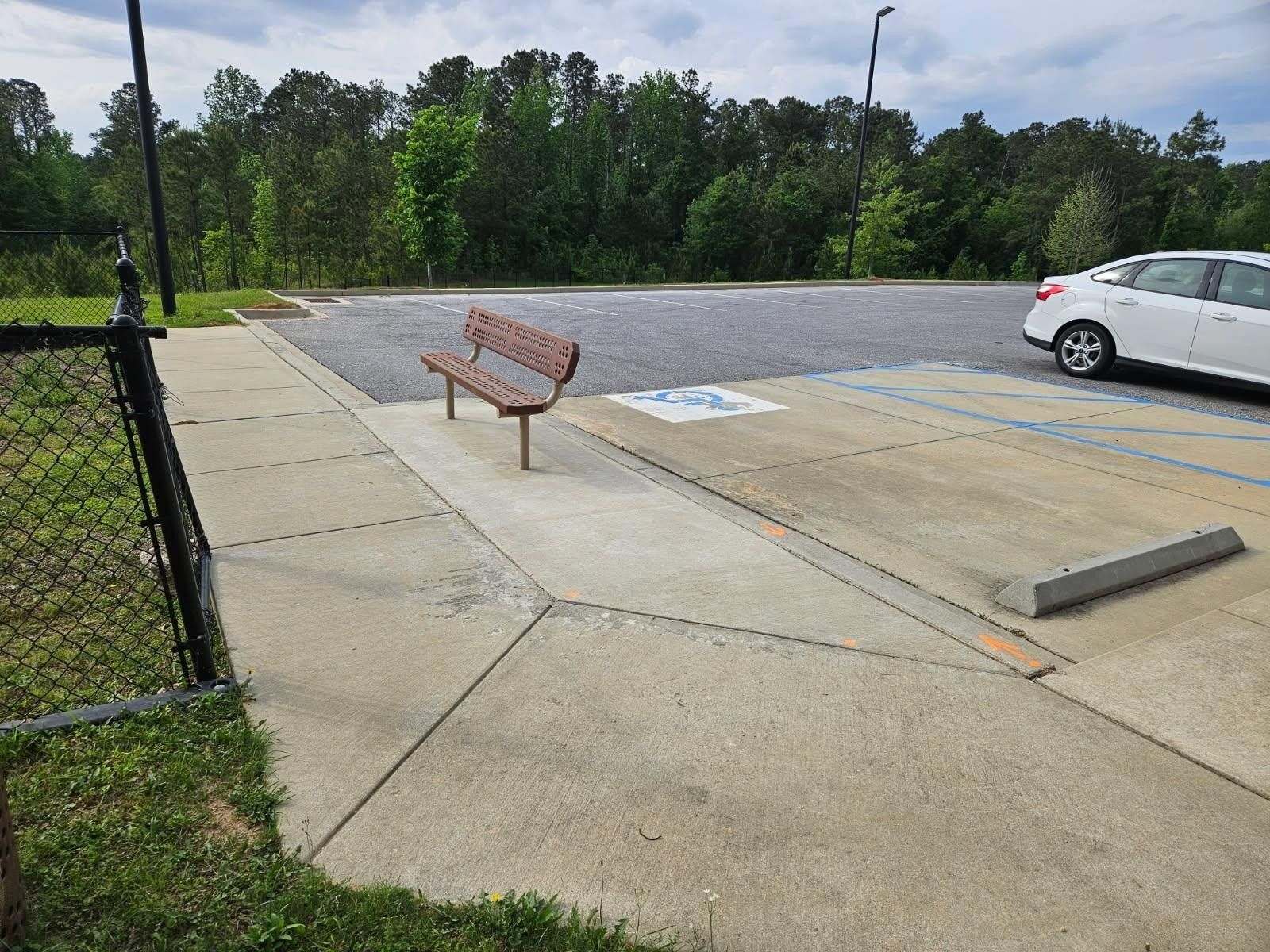 Concrete walkway with a park bench and a disabled parking space in a parking lot.