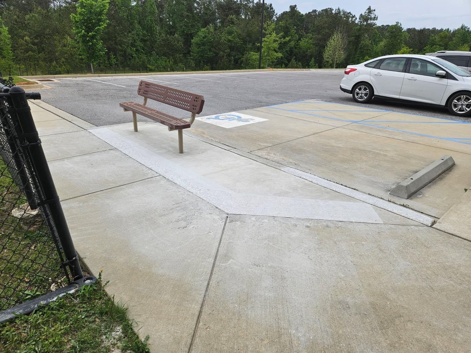 Concrete path with bench near accessible parking space, leading to asphalt lot; white car parked.