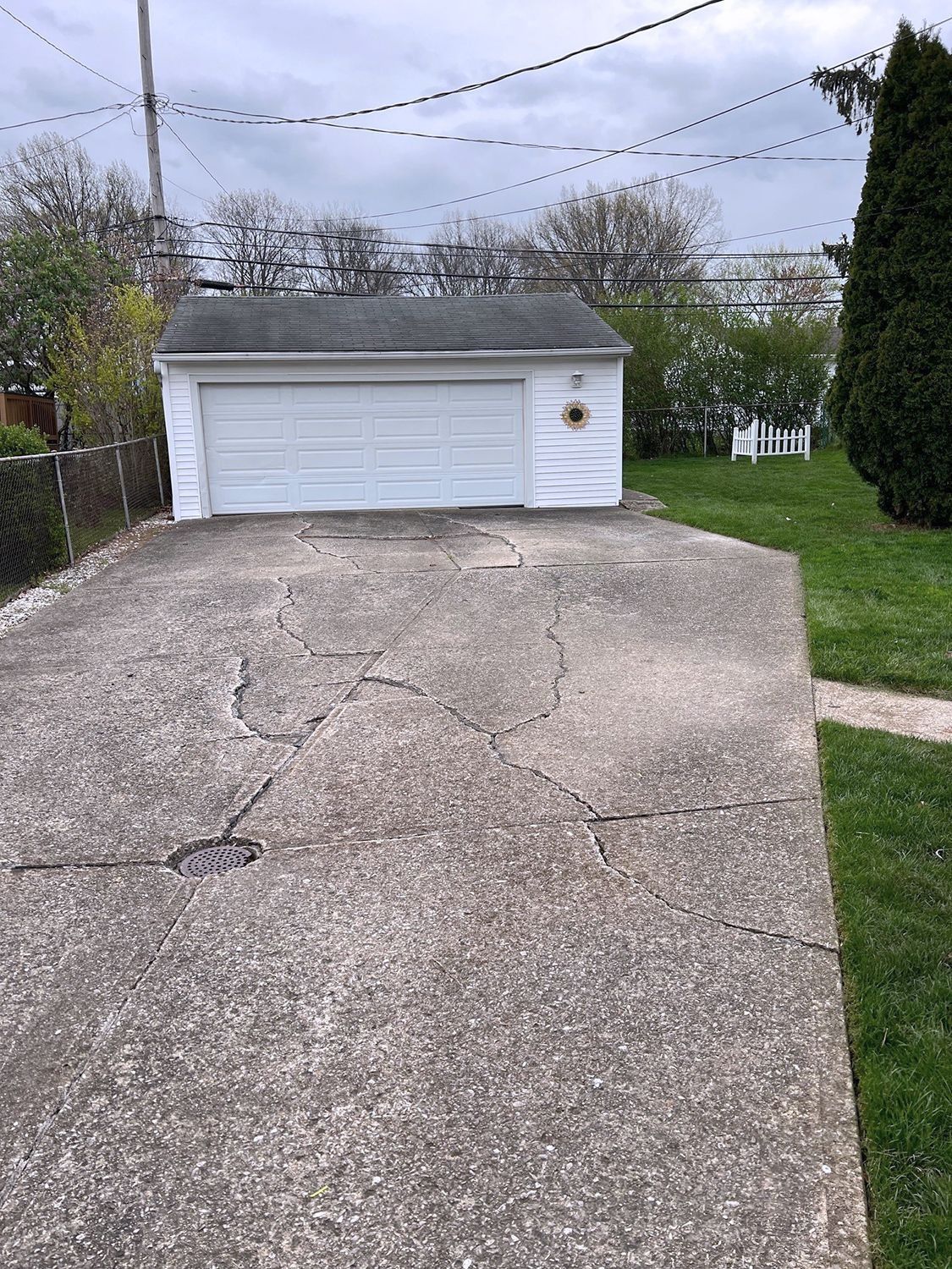Cracked concrete driveway leading to a white garage with closed door; green grass and trees in background.
