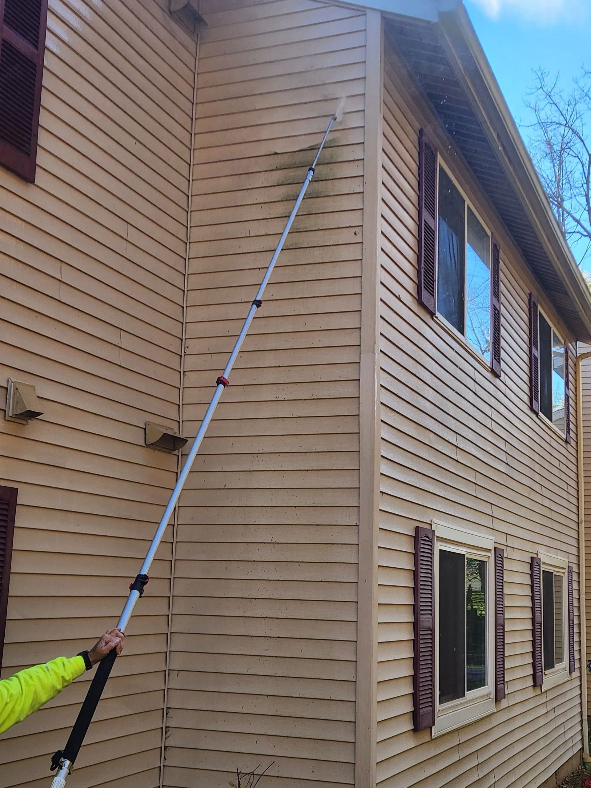 Person washing beige siding with a long-handled brush on a two-story building; brown shutters.