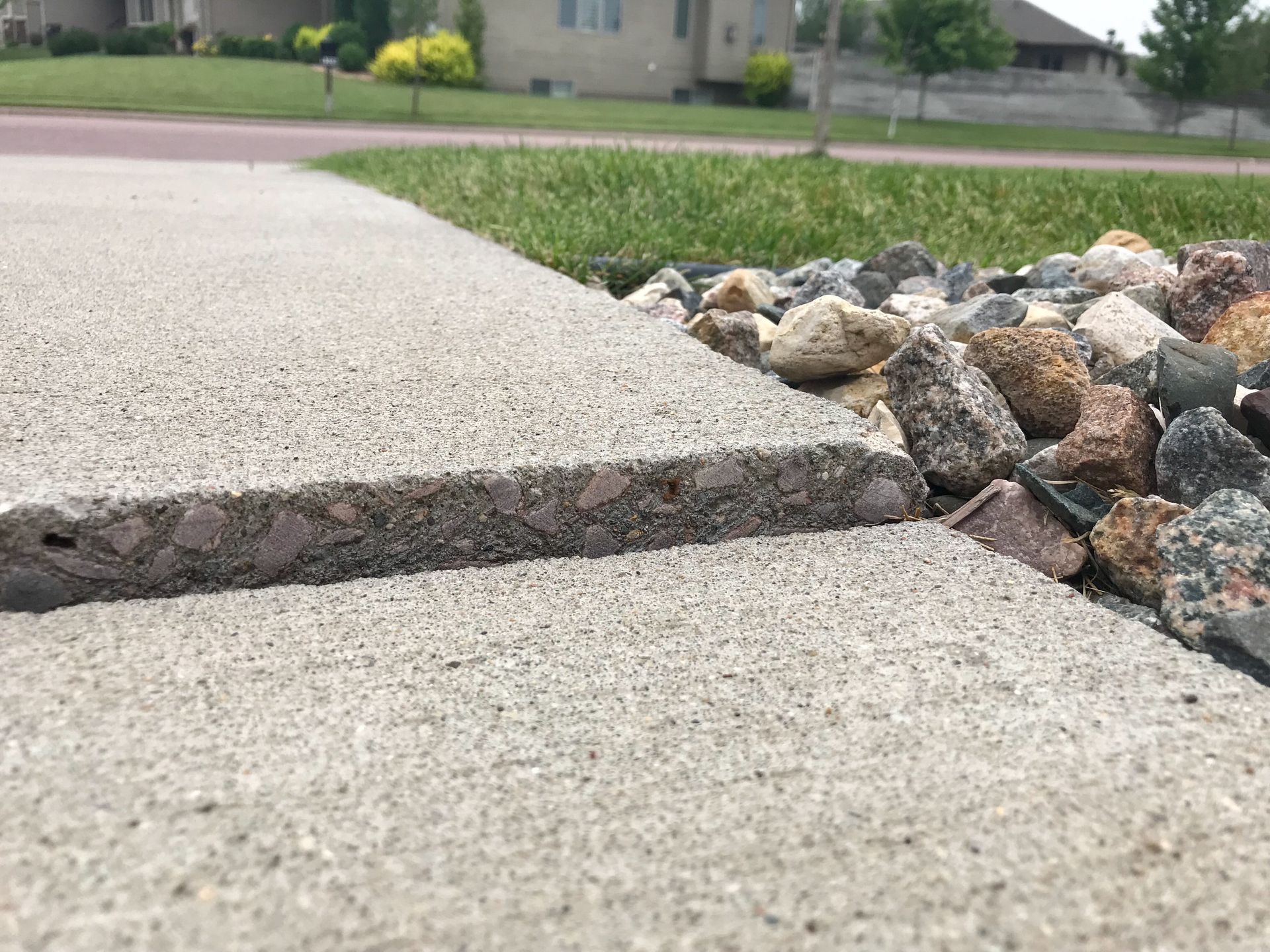 Close-up of a concrete sidewalk's edge with exposed aggregate, bordering a rock bed and green grass.