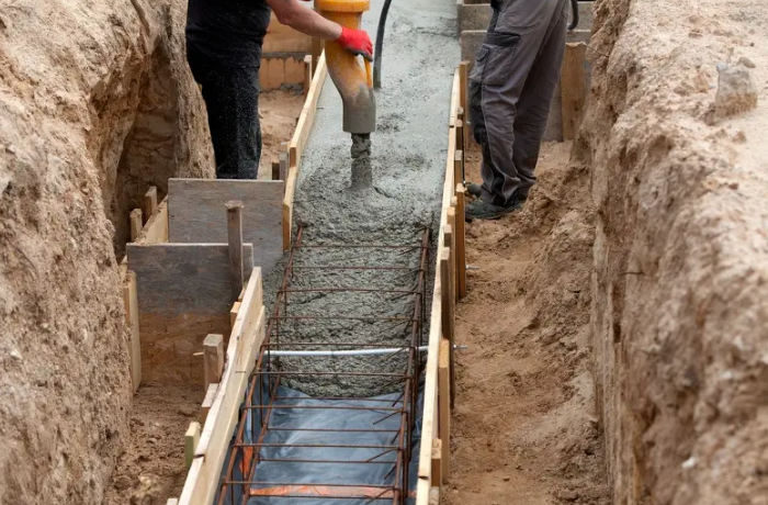Workers pouring concrete into a trench foundation with wooden forms and rebar.
