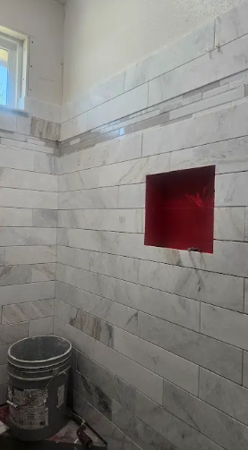 A tiled shower wall in progress, featuring white marble tiles and a rectangular recessed niche with a red waterproof base.
