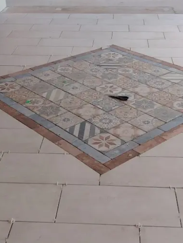 A decorative square of patterned tiles set into a floor of large, plain light-colored rectangular tiles.