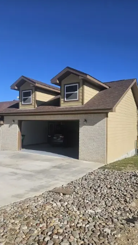 A two-story garage with light-colored siding and stone-textured front facade, featuring two dormer windows under a blue sky.