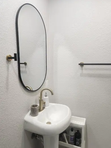 A pedestal sink with a brass faucet and an oval, black-framed mirror mounted on a white textured wall.