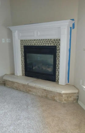 A white fireplace mantel surrounds a black firebox with tiled interior, sitting on a raised stone hearth in a room.
