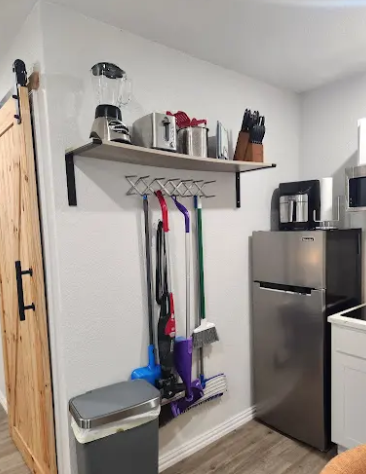 A wooden kitchen shelf holds small appliances, with cleaning tools hanging underneath next to a refrigerator and trash can.