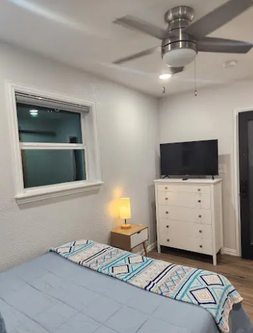 A cozy bedroom with a blue bed, a white dresser topped with a TV, a bedside lamp, and a ceiling fan.