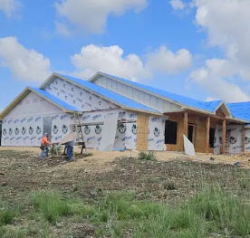 Construction workers install siding on the exterior of a house under construction with a blue roof under a sunny sky.