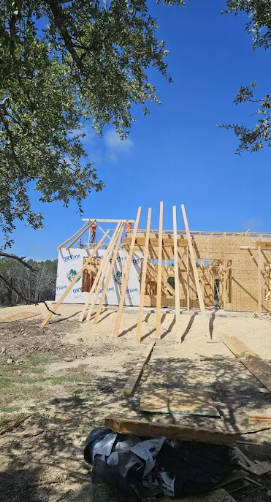 A building under construction with exposed wooden framing against a clear blue sky, framed by tree branches.