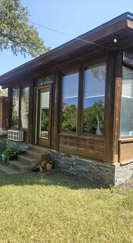 A wooden, sunroom-style building with large windows and a stone foundation base, situated on a grassy lawn.